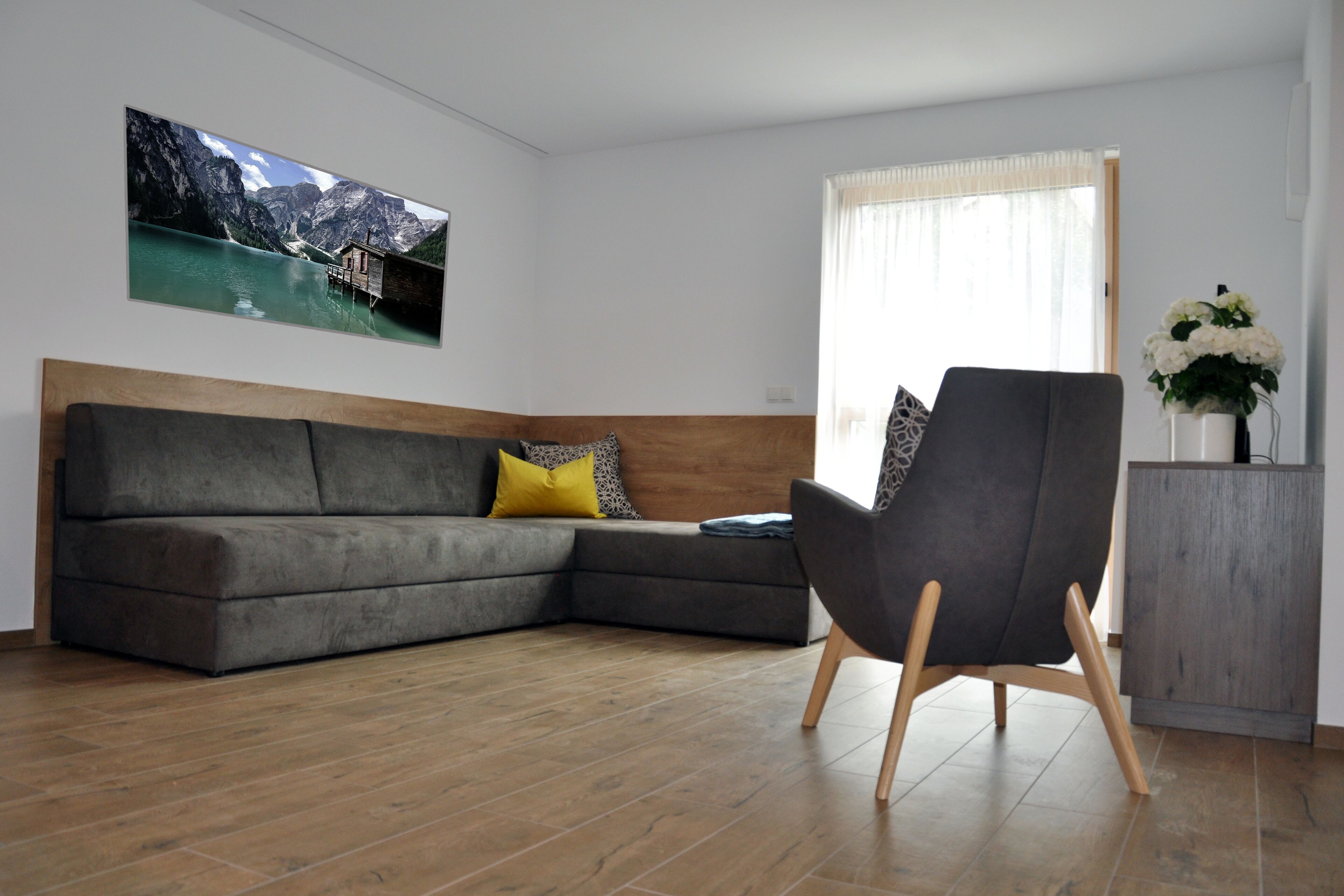 A modern living room with a grey sectional sofa, a wooden headboard, a dark armchair, and a wooden cabinet.