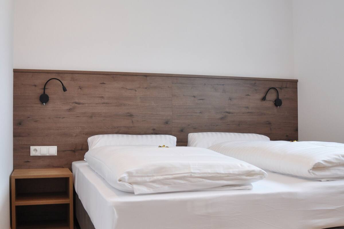 A bedroom with a wooden headboard, two white beds, a small wooden nightstand, and a wall-mounted lamp.