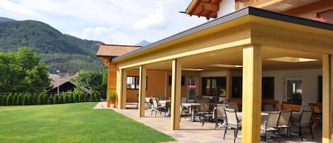 A covered outdoor seating area with tables and chairs, a well-maintained lawn, and a mountainous backdrop.