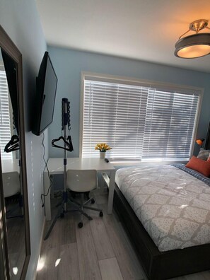 A bedroom with a bed, a desk, a chair, a TV, and a window with blinds.
