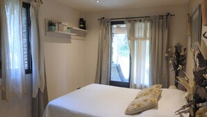 A bedroom with a bed, a window with curtains, a shelf with books and a bottle, and a view of the outside. - Stone House near Châteauneuf-du-Pape Vineyards (Bédarrides)