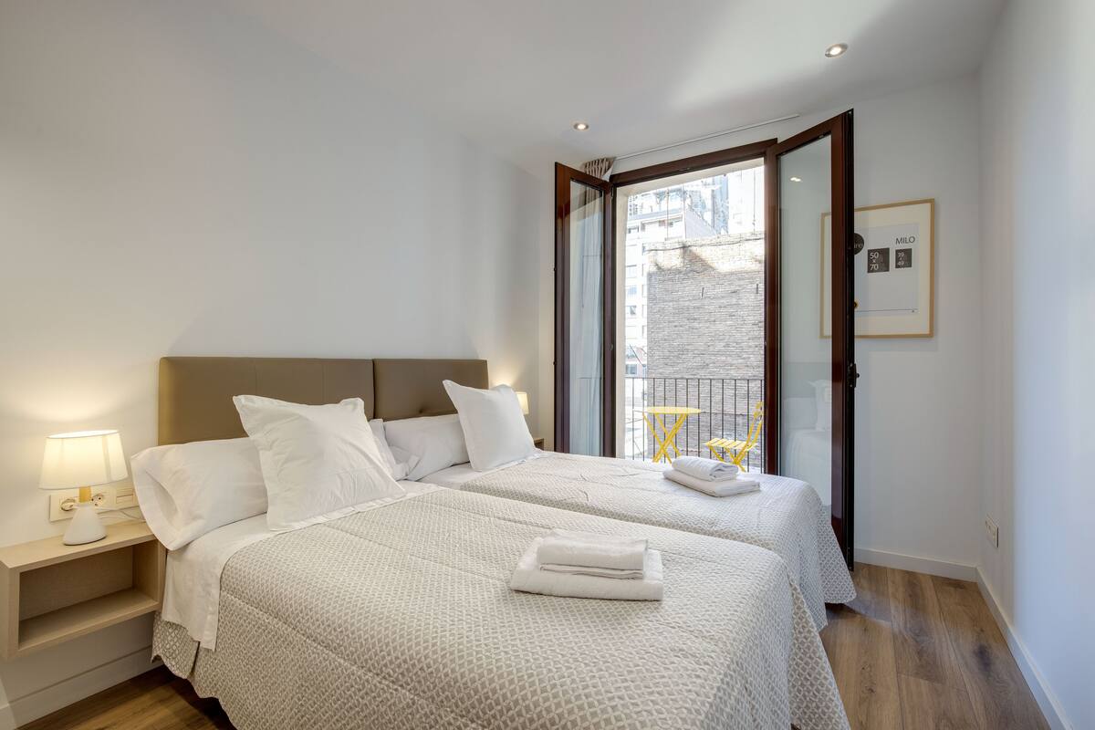 Una habitación de hotel moderna con dos camas, una mesita de noche con lámpara, un balcón con una silla amarilla y un cuadro en la pared.