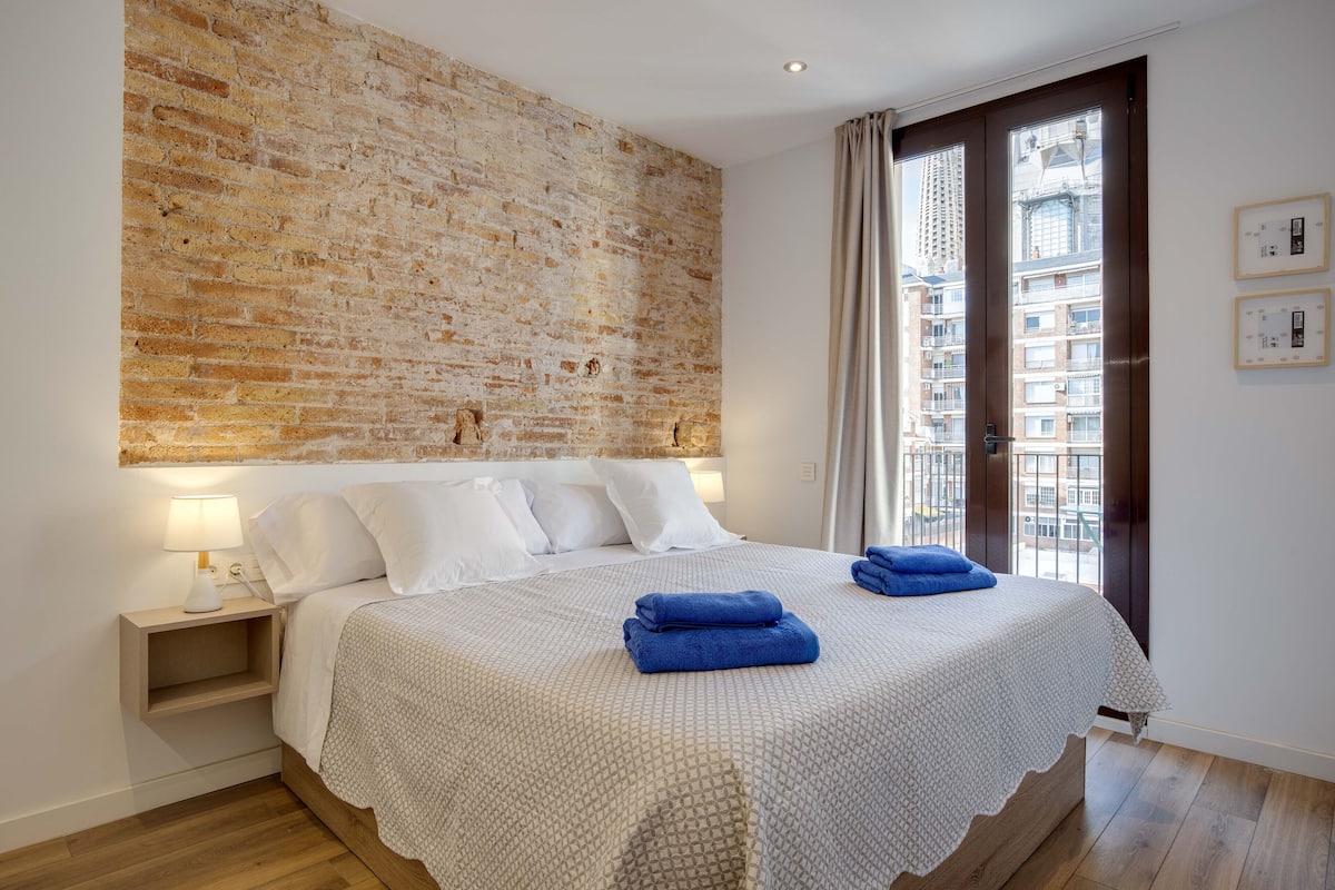 Un dormitorio con pared de ladrillo, una cama con ropa de cama blanca, una mesita de noche con lámpara y vistas a edificios a través de una ventana.