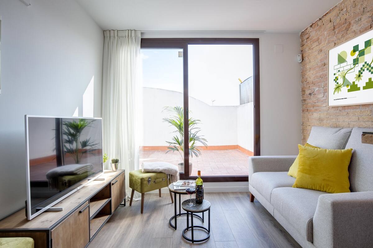 Una sala de estar moderna con un televisor de pantalla plana, un sofá con un cojín amarillo, una mesita redonda con una copa de vino y una botella, y un otoman verde.