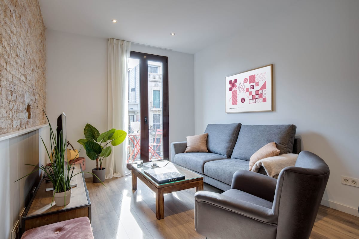 Una sala de estar moderna con un sofá gris, una mesa de centro de vidrio y un ventanal con vistas a edificios.