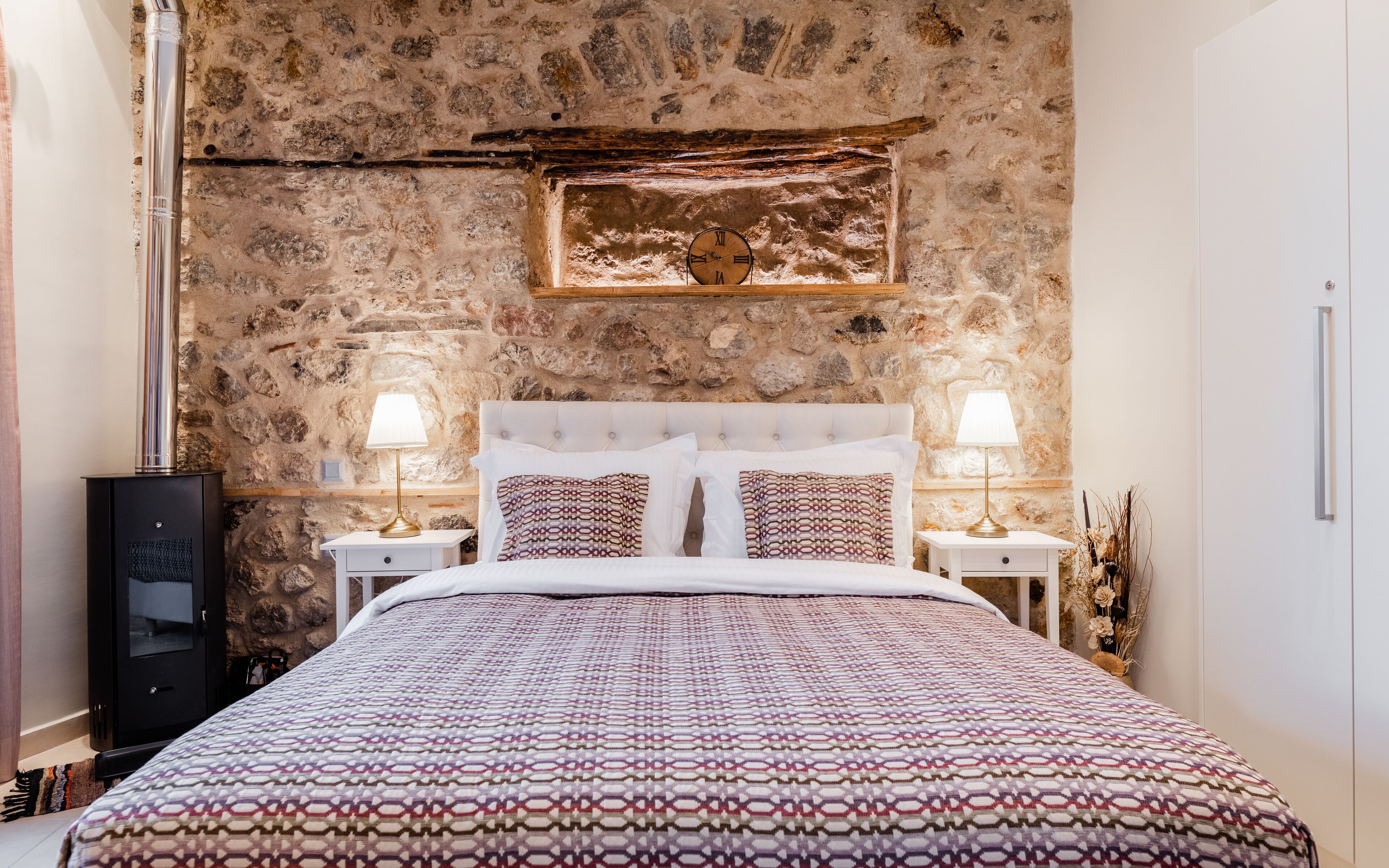 A bedroom with a stone wall, a bed with patterned bedding, two bedside tables with lamps, and a clock on the wall.
