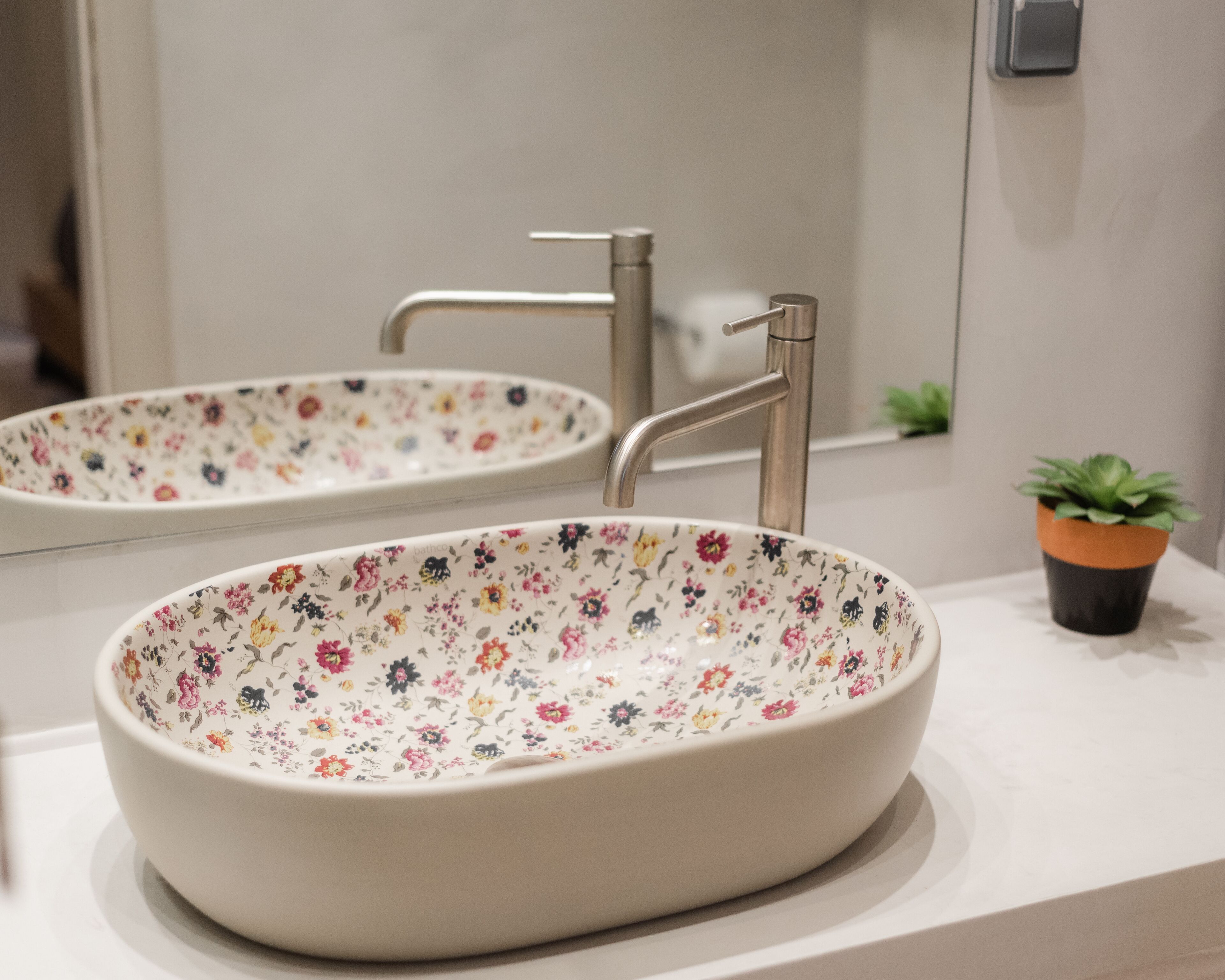 Een badkamer met een witte wastafel, een wasbak met bloemenprint en een kleine potplant op het aanrecht.