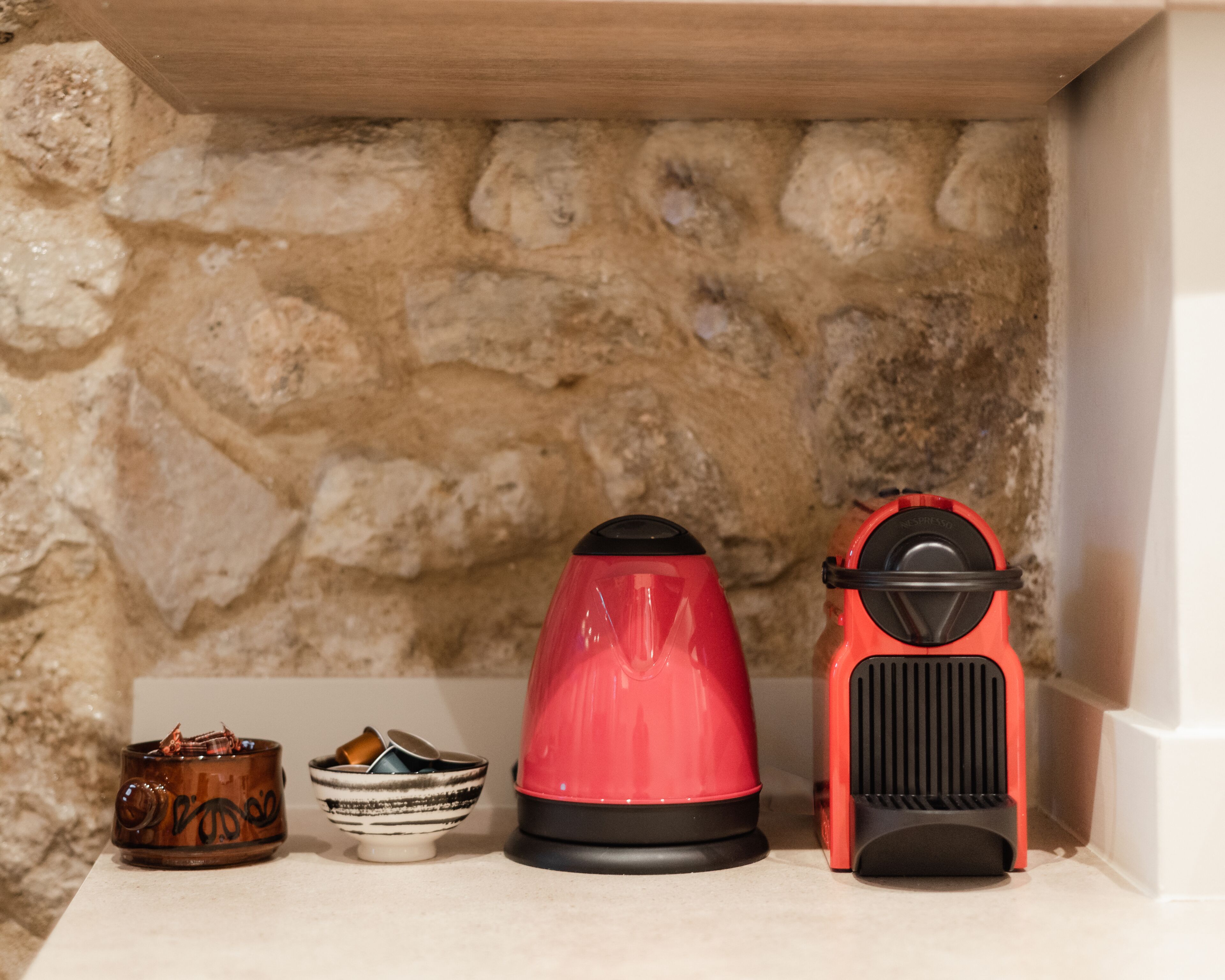A shelf with a coffee maker, a kettle, and a red appliance against a stone wall.