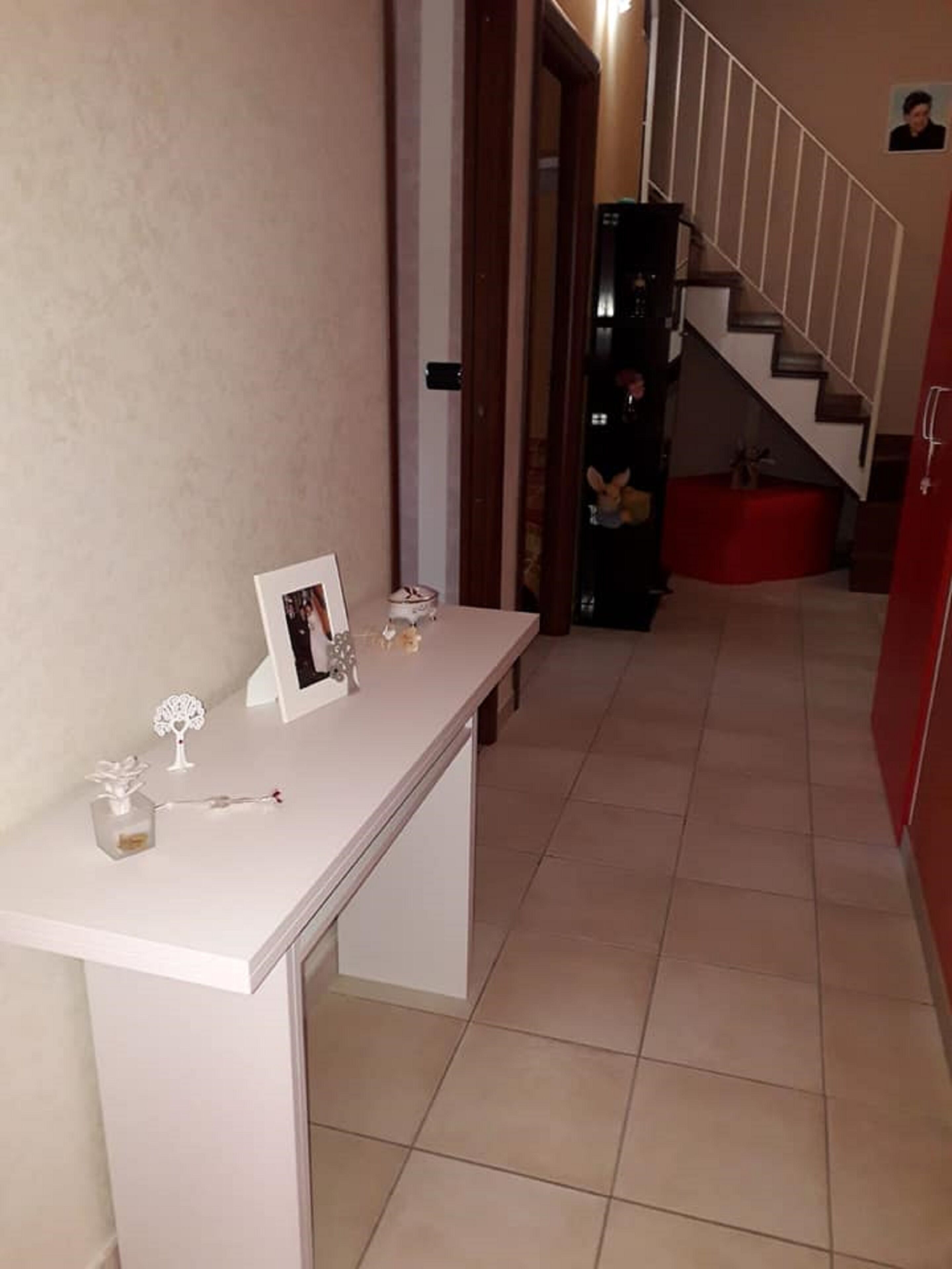 Ein Flur mit einem weißen Konsolentisch, auf dem ein gerahmtes Foto, ein kleines baumförmiges Ornament und eine Parfumflasche stehen. Im Hintergrund ist eine Treppe mit einer roten Akzentwand und einer kleinen Statue zu sehen.