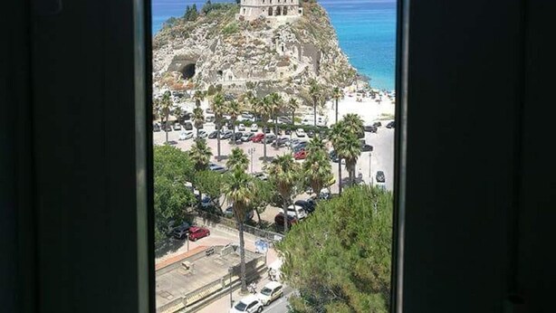 Blick aus einem Fenster auf ein historisches Gebäude auf einer Felsformation, unterhalb davon ein Strand und ein Parkplatz.
