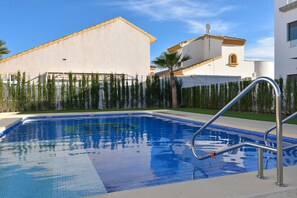 A swimming pool with a metal ladder, surrounded by a fence and palm trees, with buildings in the background.