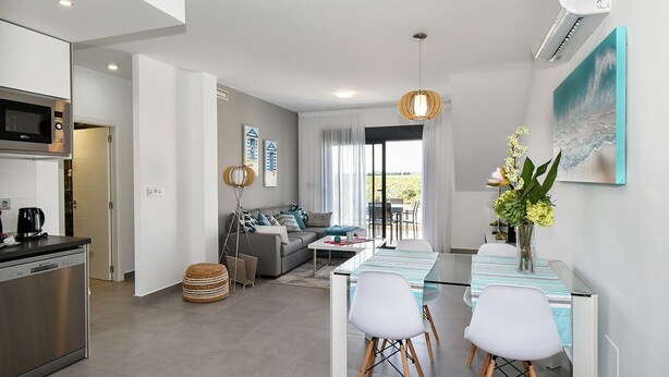 Een moderne keuken met een ingebouwde magnetron en vaatwasser, een eethoek met tafel en stoelen, een bank met kussens, en een groot raam met gordijnen.