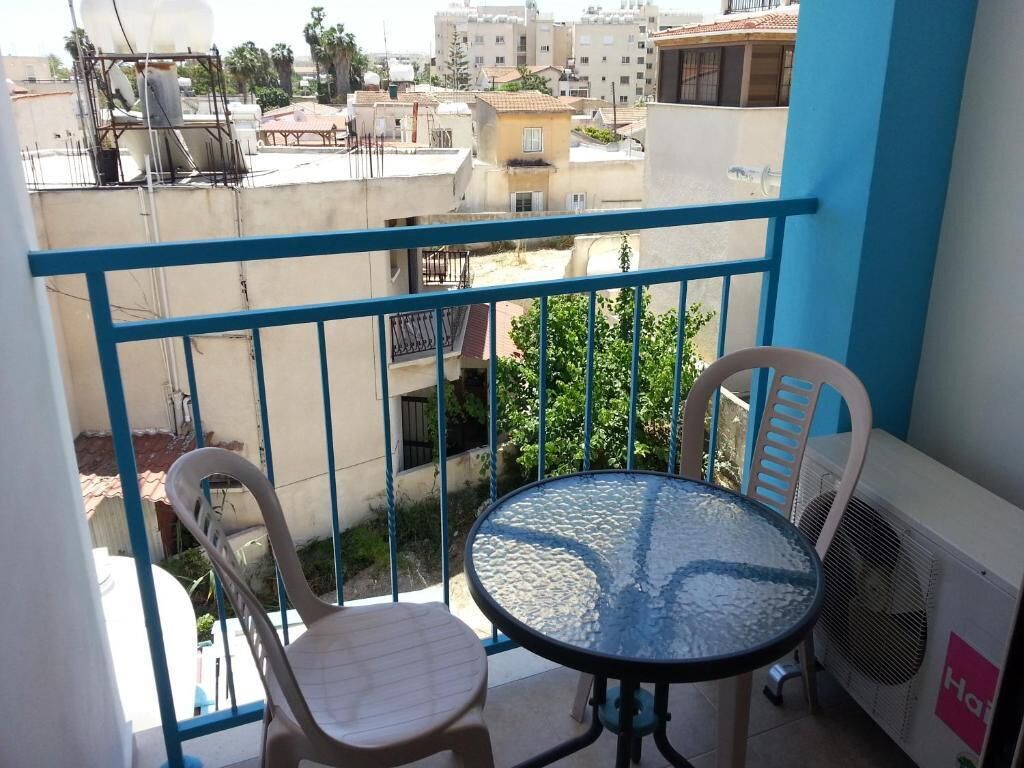 Un balcon doté d’une table ronde en verre et de deux chaises, offrant une vue sur un quartier résidentiel avec ses immeubles et sa verdure.