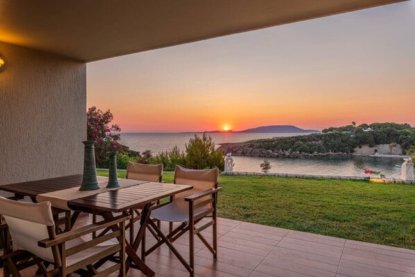 A terrace with a table and chairs overlooking a sunset over a body of water, with greenery and a distant hill.
