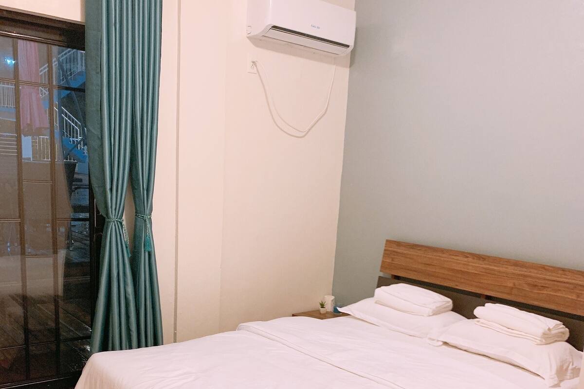 A hotel room with a bed, a wooden headboard, a window with curtains, and an air conditioning unit on the wall.