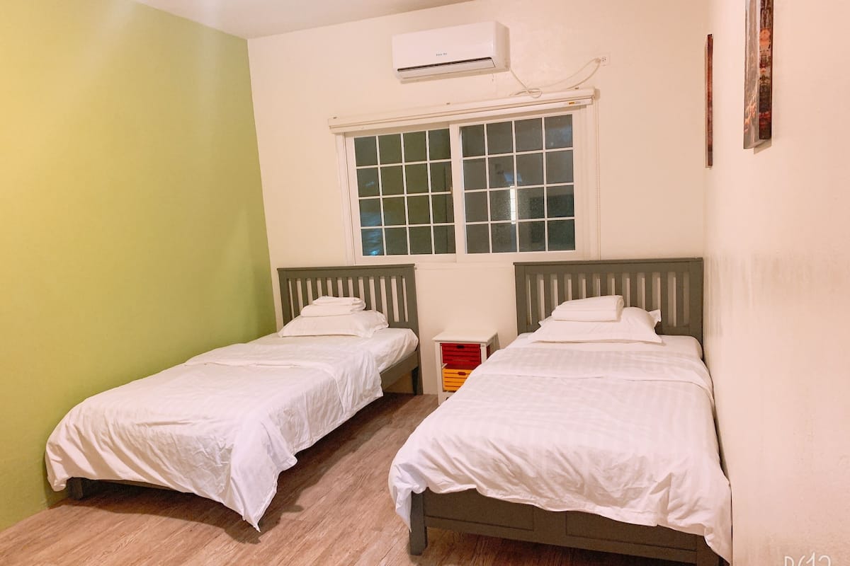 A room with two single beds, a window with blinds, an air conditioning unit, and a small storage unit with drawers.