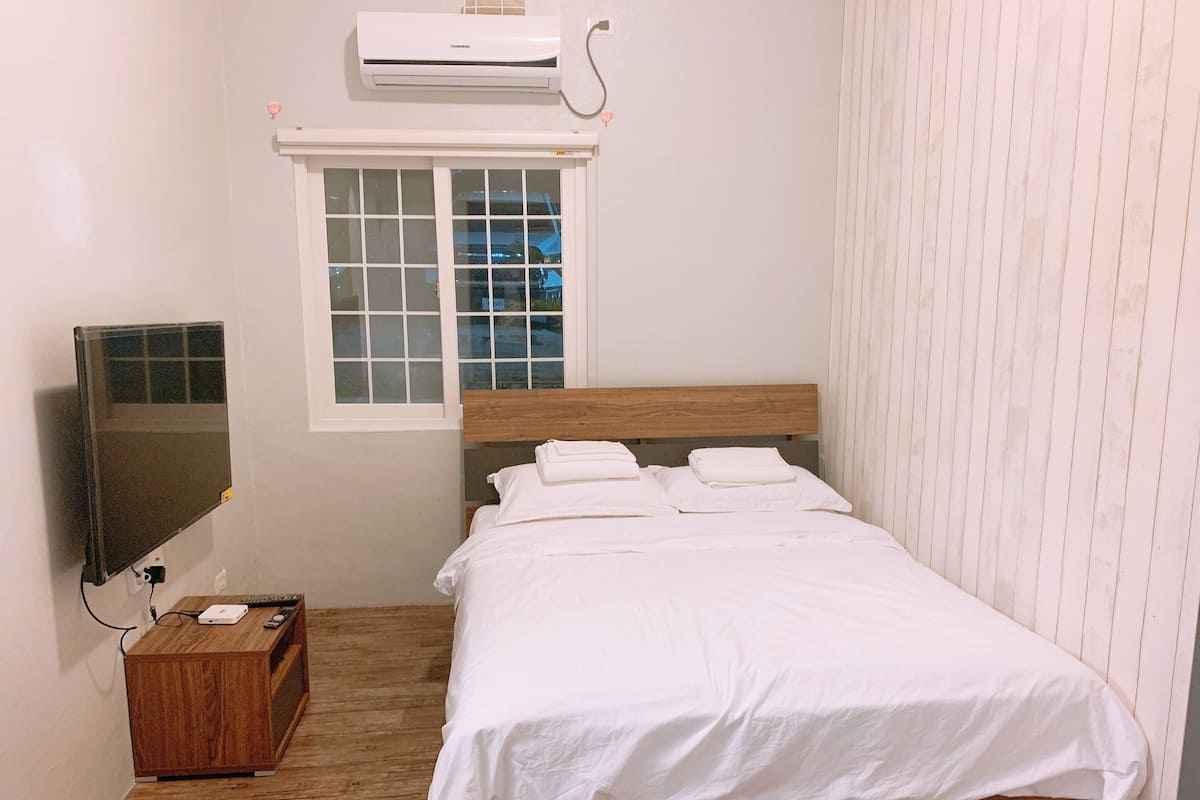 A neatly made bed with white linens, a wooden headboard, and a window with blinds.