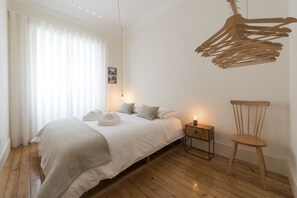 A bedroom with a bed, a wooden floor, a nightstand, a chair, and a hanging light fixture.