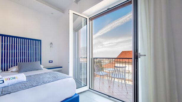 A modern bedroom with a large bed, a blue headboard, and a view of the outdoor balcony with chairs. - Al Campanile (Sant'Agnello)