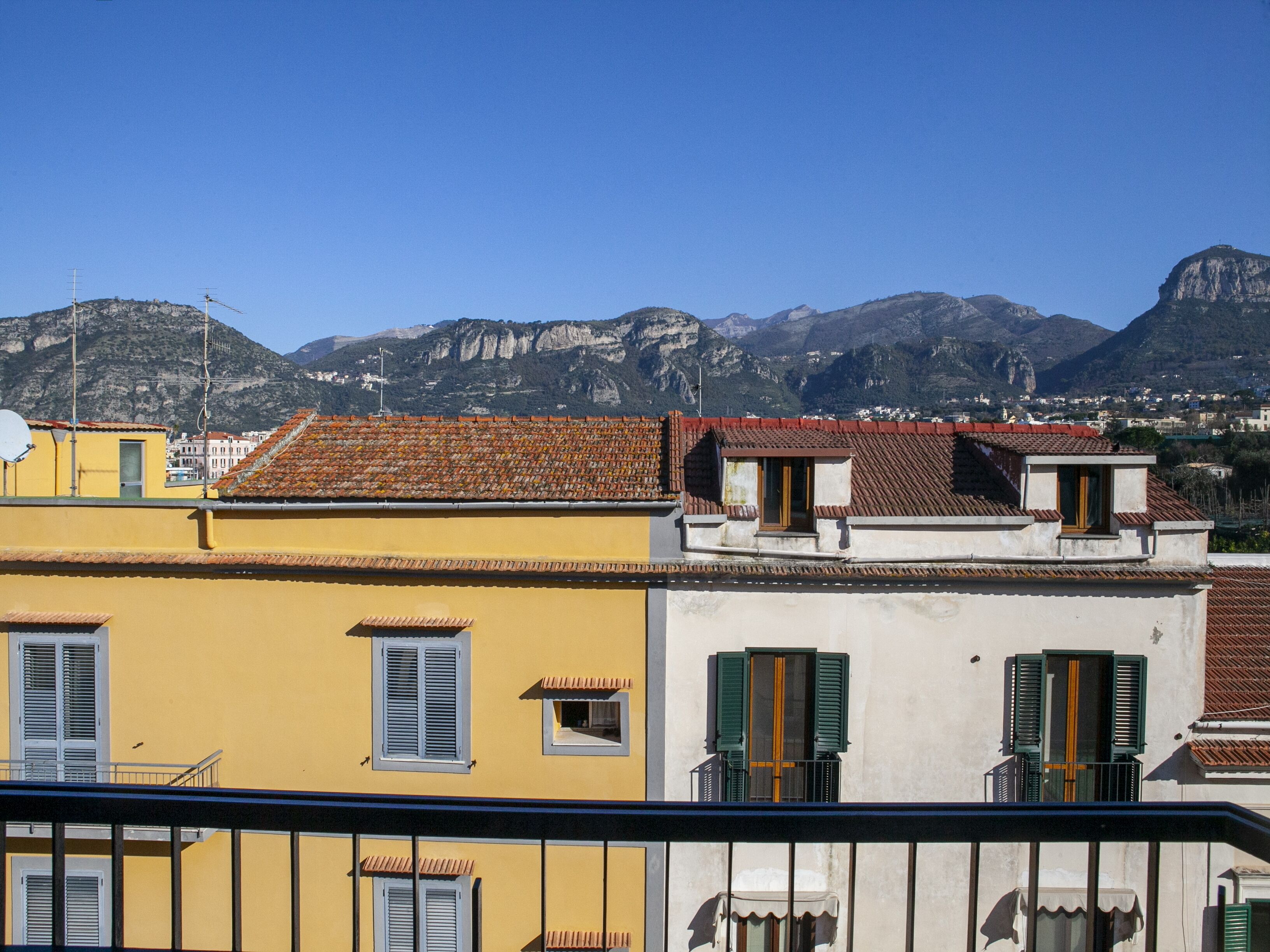 A row of buildings with red-tiled roofs and balconies, set against a backdrop of mountains.