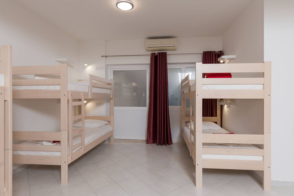 A room with three bunk beds, a window with a curtain, an air conditioning unit, and a small shelf with a lamp.
