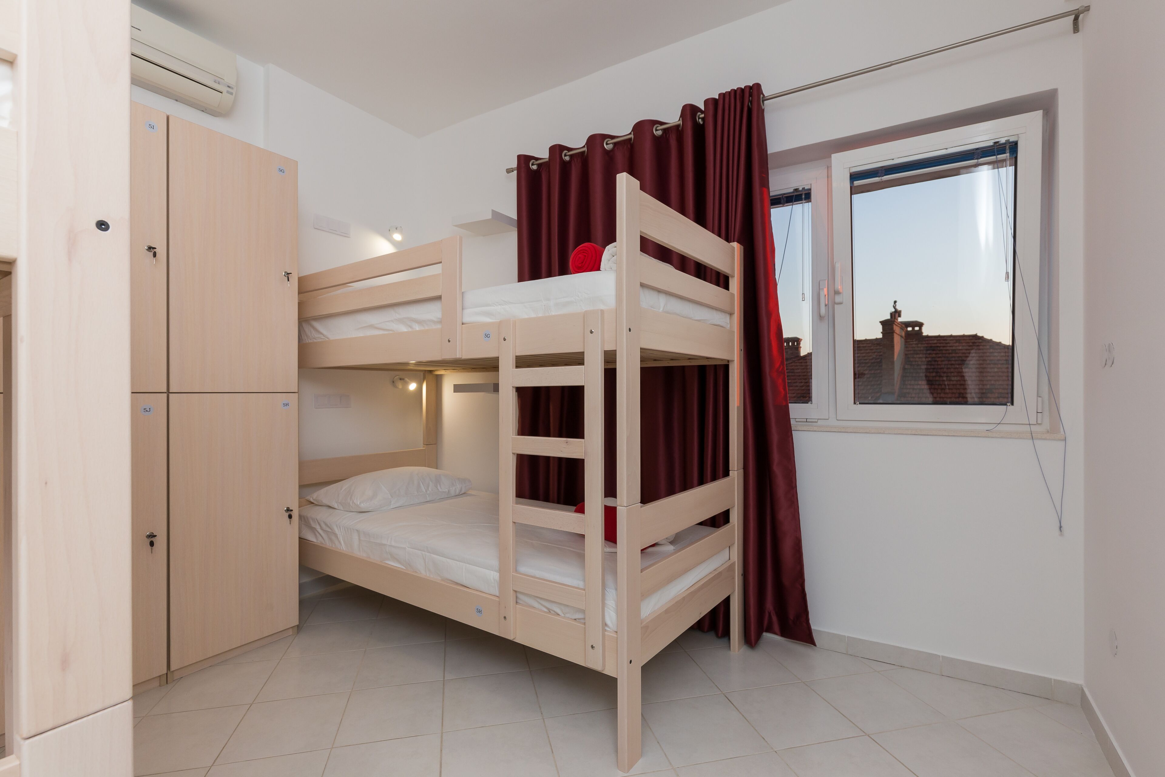 A bunk bed room with a wardrobe, a window with curtains, and a view of buildings outside.