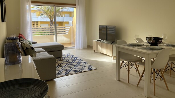 Una sala de estar moderna con un sofá, una mesa de comedor para dos personas, un televisor y vista a un edificio amarillo a través de un ventanal grande.