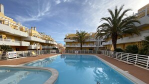 Une piscine entourée d’une clôture blanche, avec un palmier et des bâtiments jaunes en arrière-plan.