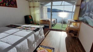 A bedroom with a bed, a small table, a chair, a window with a view of the sea, and a painting on the wall.