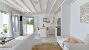 A modern, minimalist living room with a sofa, a dining table, and a kitchen area visible through an open door. - Mythology Naxos Villas & Suites (Naxos)