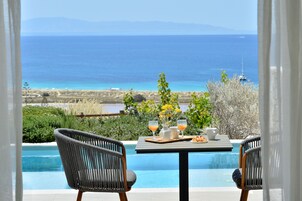 Mesa à beira da piscina, com bebidas e um prato, com vista para o mar azul e as montanhas ao longe.