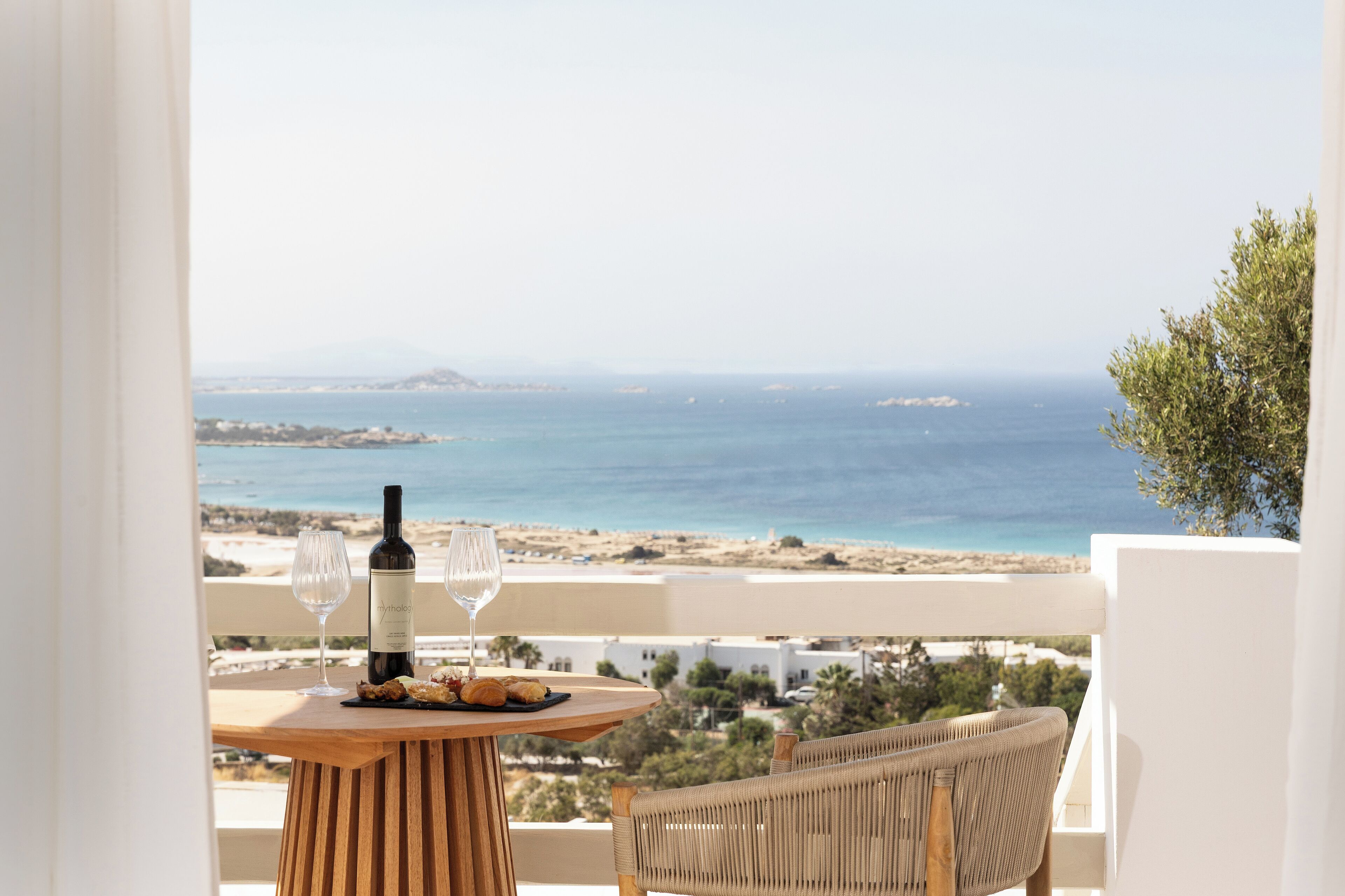 A balcony with a table set for two, a bottle of wine, and a glass, overlooking a coastal landscape with buildings and vegetation.