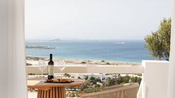 A balcony with a table set for two, a bottle of wine, and a glass, overlooking a coastal landscape with buildings and vegetation.