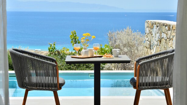 A dining table with a view of the ocean, two wicker chairs, and a stone wall. - Mythology Naxos Villas & Suites (Naxos)