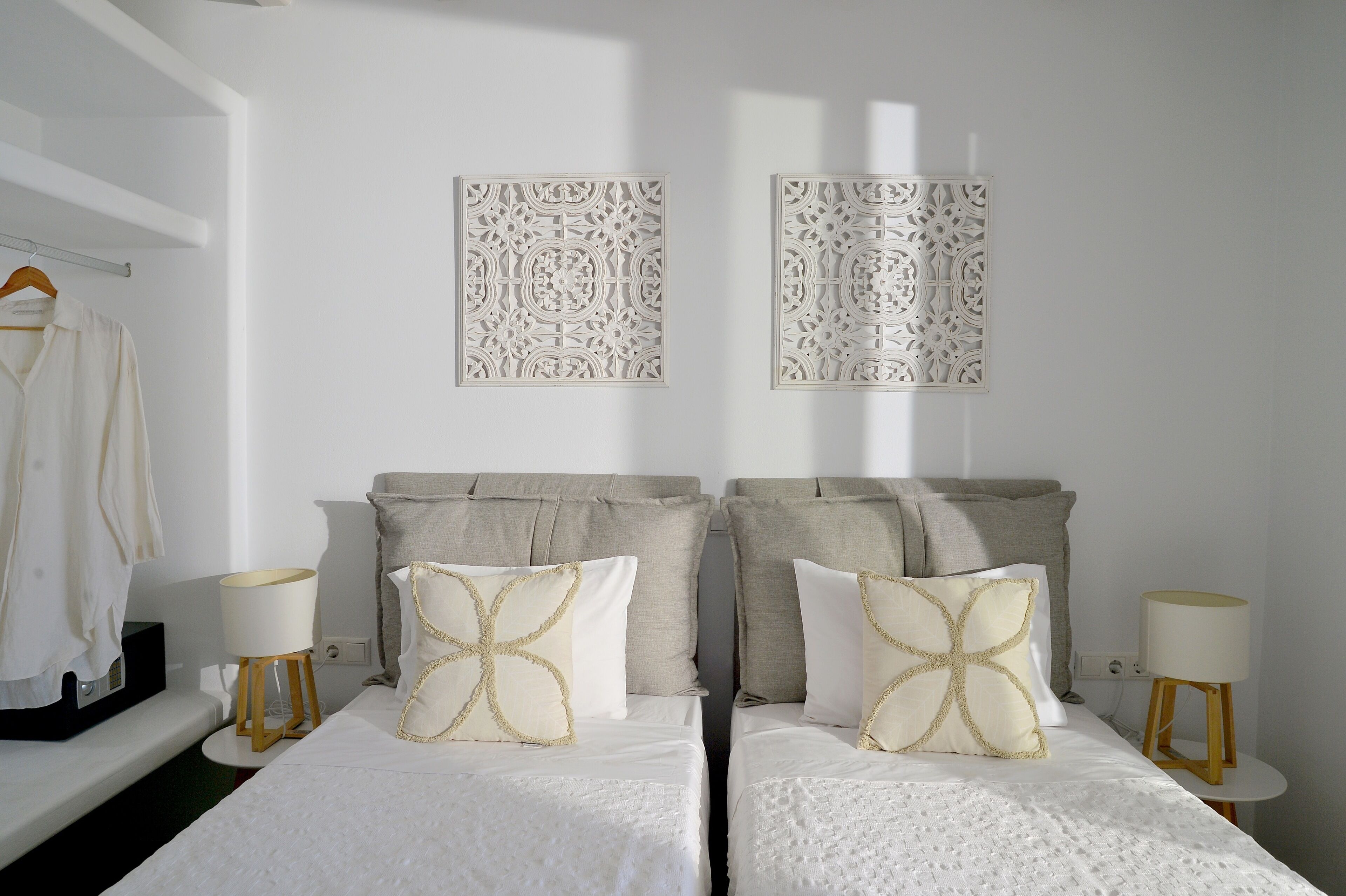 Een slaapkamer met twee bedden, elk voorzien van een witte spreidek en decoratieve kussens, een nachtkastje met een lamp en een wandplank met een hangende kledingstuk.