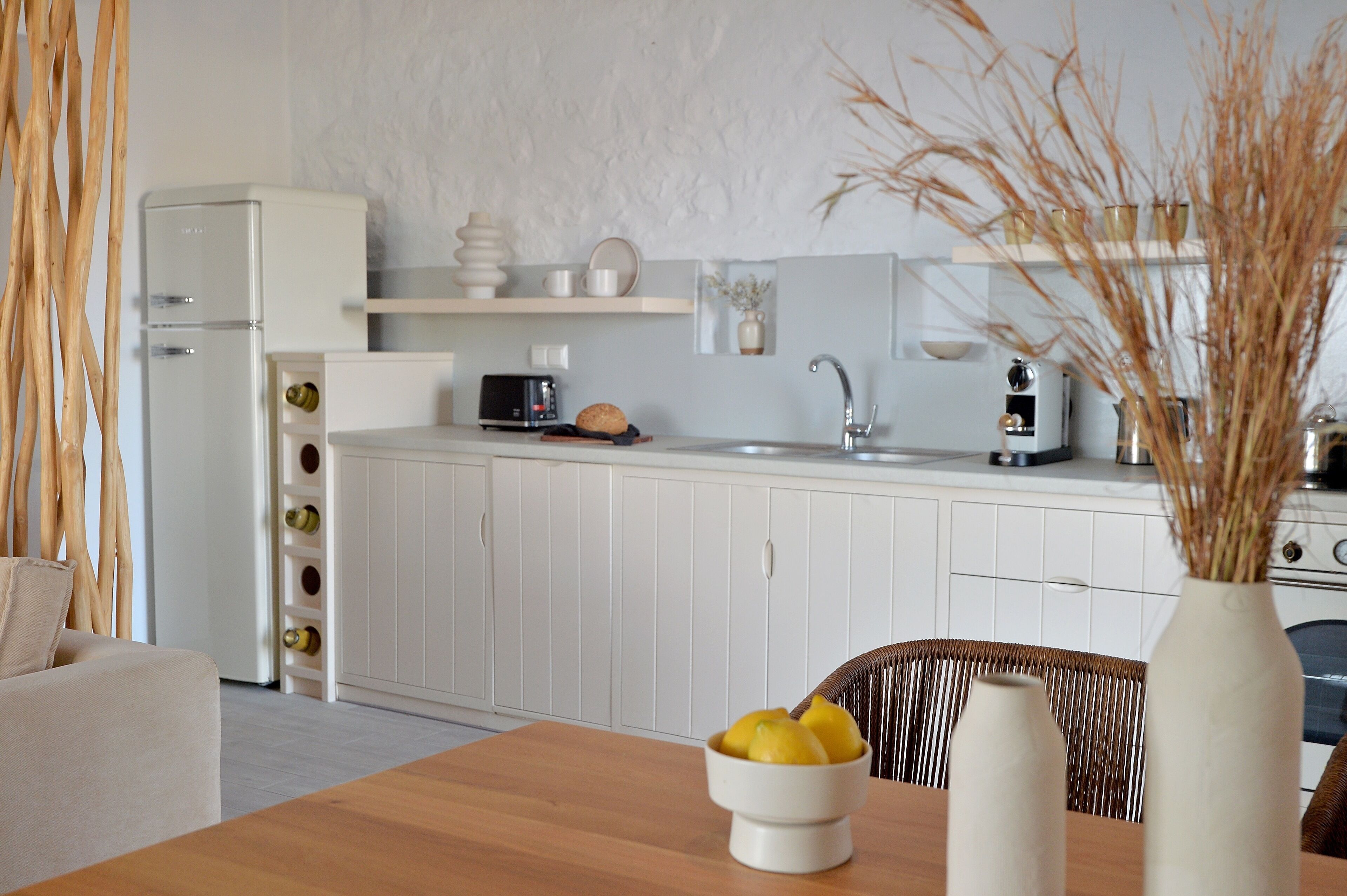 A modern kitchen with white cabinetry, a wooden dining table with a bowl of fruit, and a vase with dried plants.