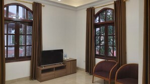 A room with a flat-screen TV on a wooden stand, two wooden chairs, and large windows with curtains. - Van Xuan Royal Hotel (Ninh Binh)