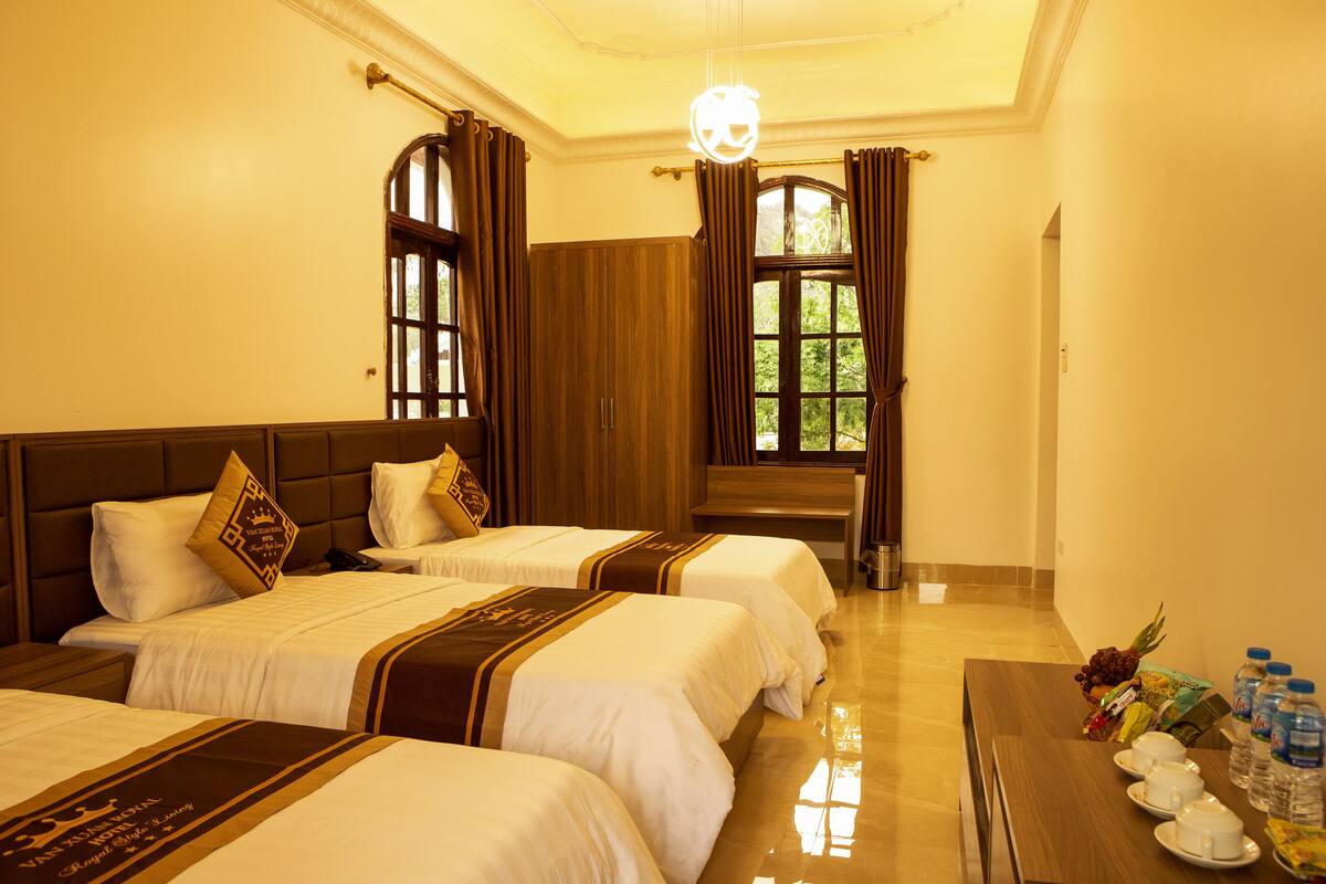 A hotel room with two beds, a wooden headboard, a dining table with plates and bottled water, and a view of greenery through the window.