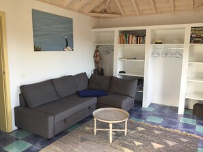 A living room with a grey sofa, a small round table, a blue pillow, and a bookshelf with books and hangers.