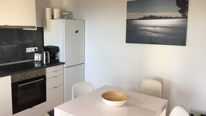 Une cuisine compacte équipée d’un réfrigérateur, d’un four et d’une machine à café. On y trouve une table à manger avec un saladier et des chaises. Un tableau représentant un paysage pittoresque, avec un lac et des montagnes, est accroché au mur.