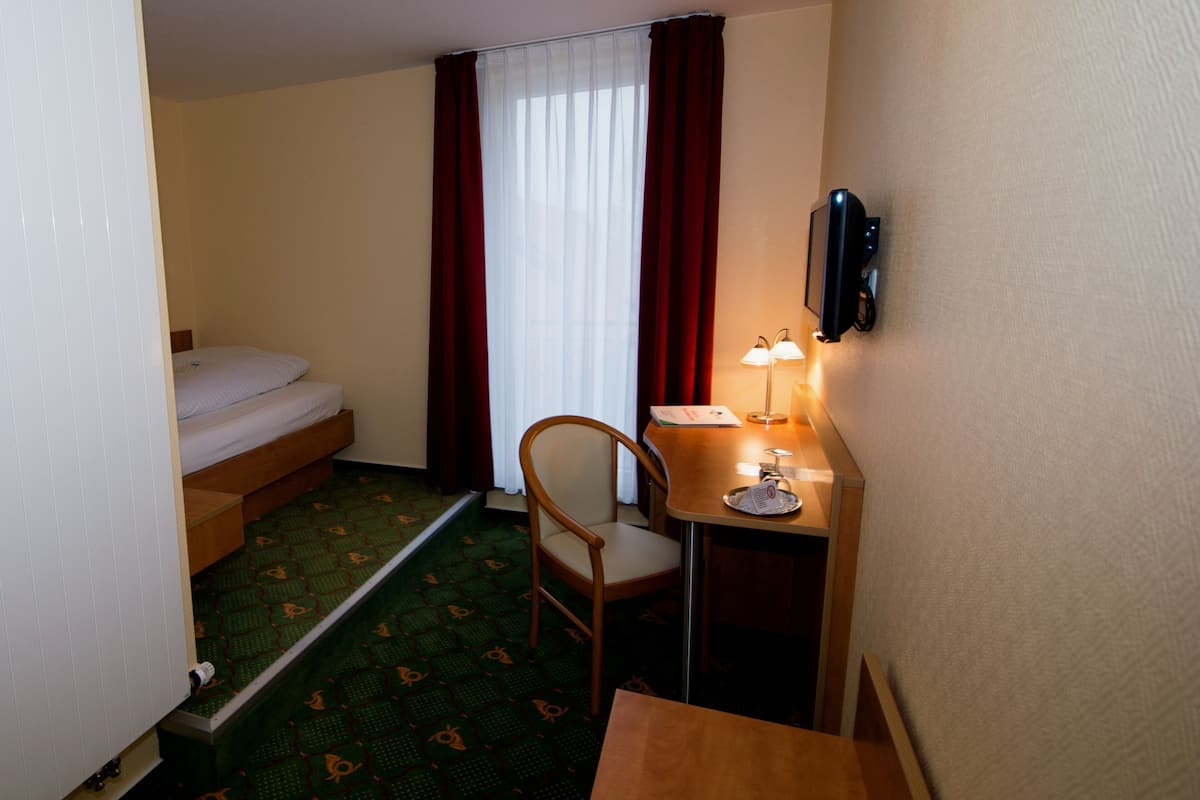 A small hotel room with a bed, a desk with a lamp, a chair, and a TV on the wall.