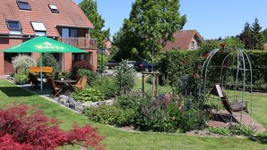 A garden area with a table and chairs under a green umbrella, a metal pergola with flowers, and a brick building in the background.