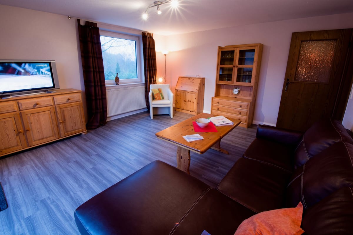 A living room with a sofa, a television, wooden cabinets, a dining table, and a window with curtains.