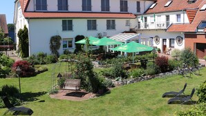 A two-story building with a red-tiled roof, a garden with a bench, and outdoor seating with umbrellas.