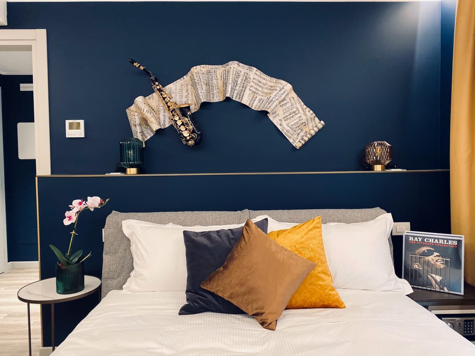 A bedroom with a dark blue wall, a saxophone, and a book titled "Ray Charles: The Ultimate Collection" on a nightstand.