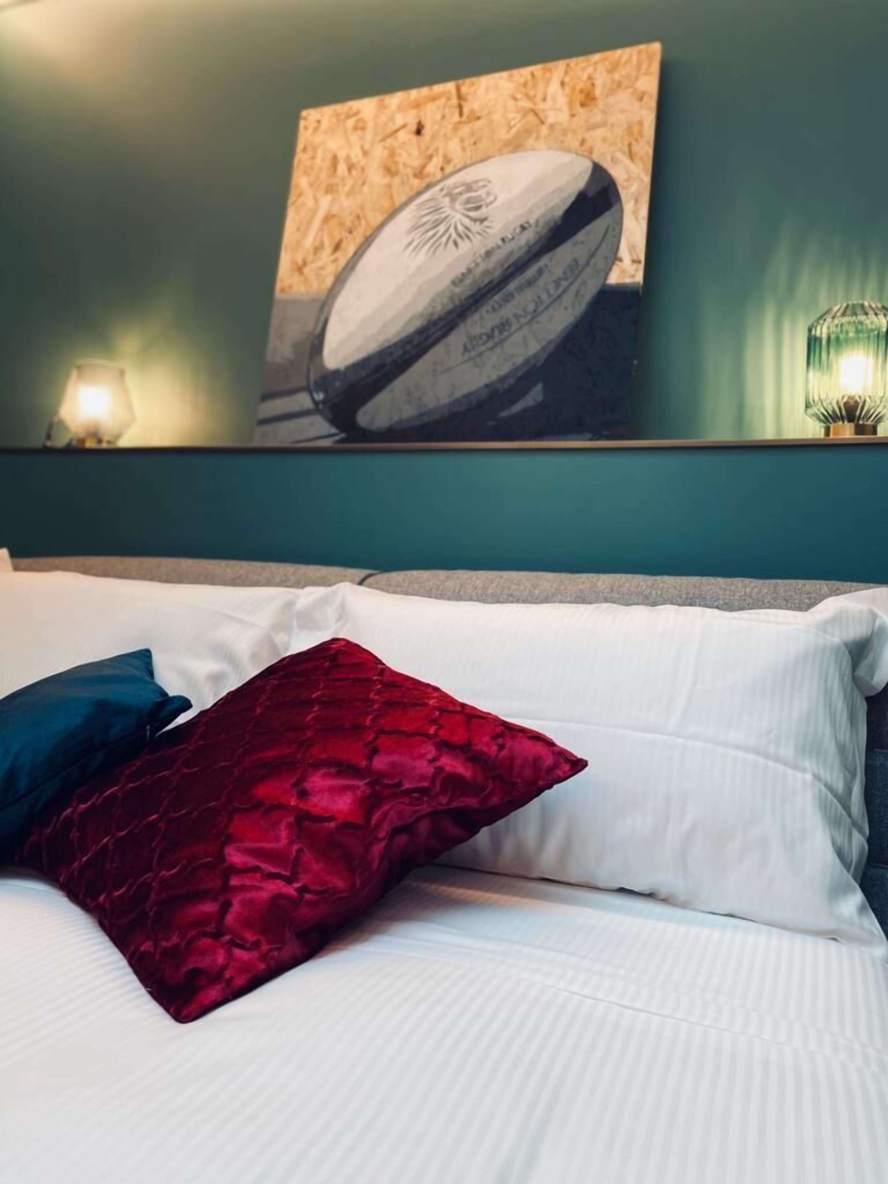 A bed with white and red pillows, a teal wall, and a framed picture of a rugby ball.