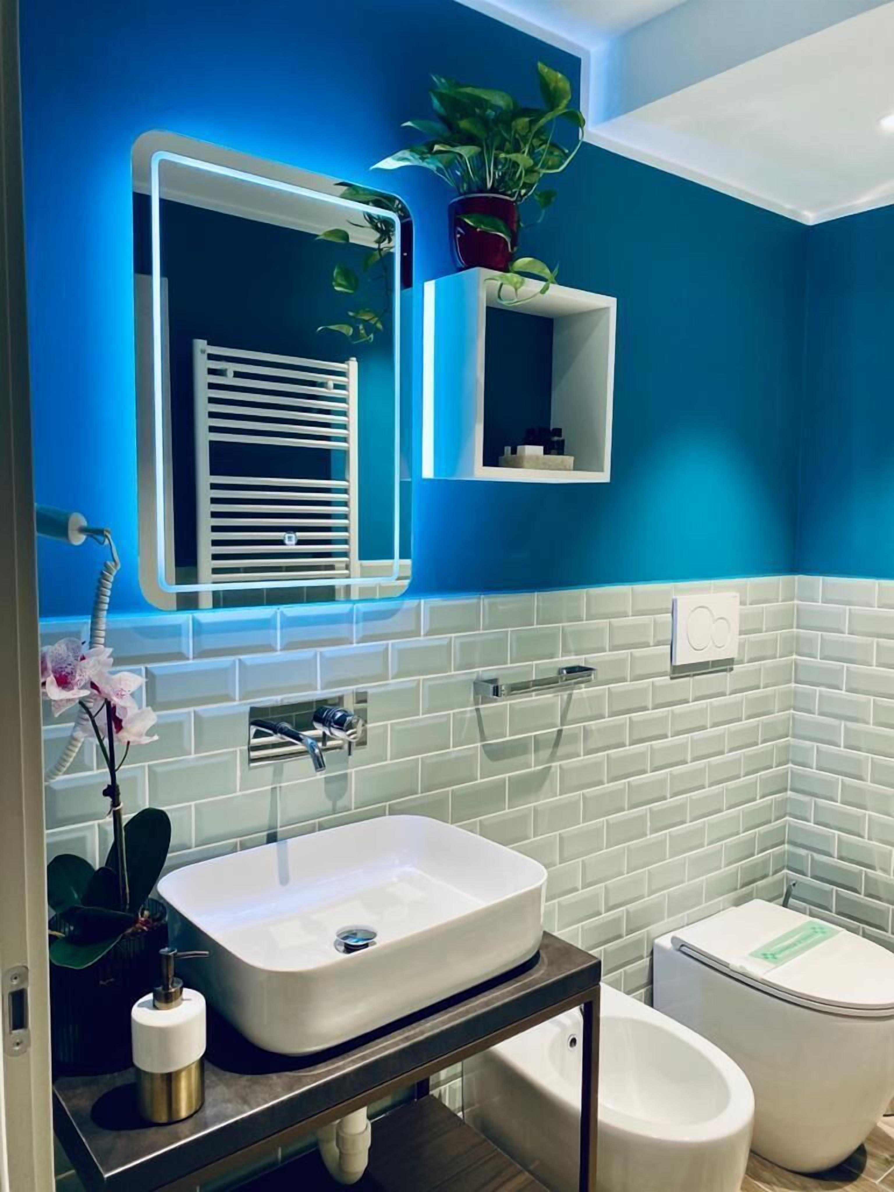 A modern bathroom with a blue accent wall, a white sink, a toilet, and a mirror with a lighted frame.