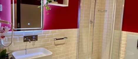 A bathroom with a white sink, a bidet, a glass-enclosed shower, and a mirror with a lighted frame.