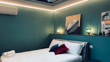 Una cama bien hecha con sábanas blancas y un cojín rojo, un aire acondicionado de pared y una obra de arte enmarcada sobre la cama.