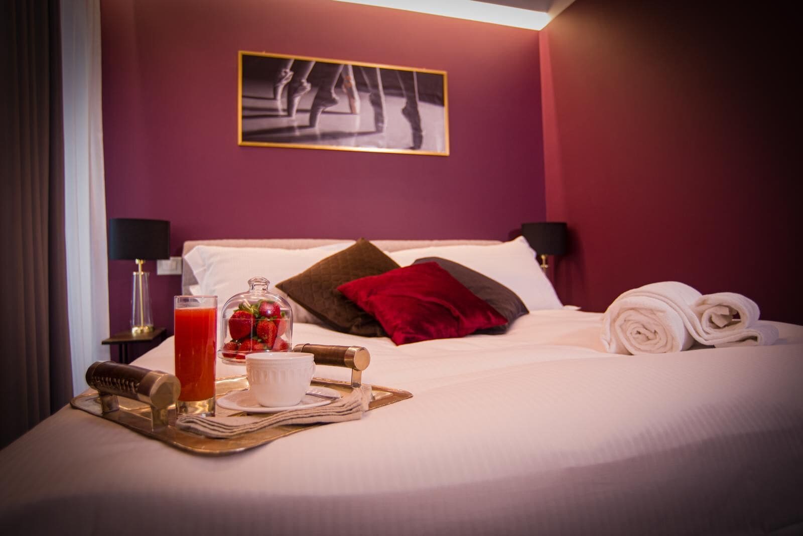 A neatly made bed with a red pillow, a glass of juice, a bowl of strawberries, and rolled towels on a tray.